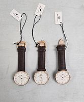 Front view of three Brigada watches side by side, showing white dials, gold-tone cases, and dark brown leather straps with tags attached.
