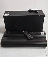 Full kit including two black soundbar units, rectangular subwoofer with visible circular speaker, remote control placed on soundbar, and power cable on top of subwoofer.