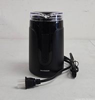 Front view of the black electric coffee grinder with clear lid and power cord coiled beside it.