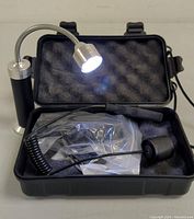 Open protective case showing the flashlight with flexible neck lit up, connected by a coiled cable, and foam padding inside the case.