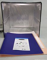 Foldable lightbox with reflective silver interior and several colored fabric backdrops beside it including blue, peach, and white.