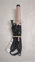 Front view of Abody hair curler showing black ergonomic handle with buttons and attached power cord coiled and held by rubber band, beige ceramic barrel visible.