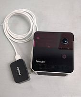 Front view of the Petcube Play 2 camera showing glossy black front with lens, sensors, and Petcube logo, connected to white USB cable and black power adapter.