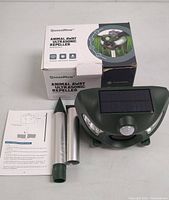Photo showing the QueenMew Ultrasonic Repeller components outside the box: the main repeller unit with solar panel, metal stake parts, instruction manual, and box.