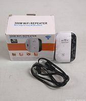 Front view of WiFi repeater device, its power adapter and USB cable next to the box showing product name and features.