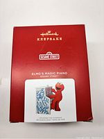Red Hallmark Keepsake ornament box with Sesame Street branding and image of Elmo playing piano ornament.