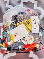 Plastic bag filled with assorted LEGO bricks and pieces including a yellow gate panel, white wheel part, gray base plate, red bricks, and other small components.