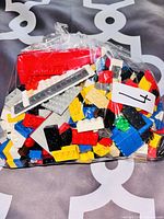 Clear sealed plastic bag with assorted LEGO and compatible building bricks in various colors and shapes, including red, white, yellow, blue, black, gray, and brown pieces.