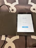 Samsung Galaxy tablet powered on displaying welcome screen connected to charger, with black protective case open beside it.