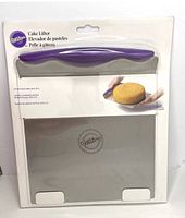 Photo of the Wilton cake lifter in original packaging showing the stainless steel lifter with purple handle and the Wilton logo, along with product information.