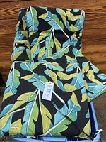 Photo showing multiple stacked cushions with green and blue leaf pattern on black, including seat and back cushions, tied with fabric loops.