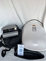 Polaroid camera with attached strap and black carrying case next to white George Foreman Lean Mean Fat Grilling Machine indoor grill