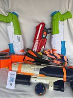 Photo shows two identical green, white, and blue Super Soaker water guns, one large white, orange, and black Super Soaker water gun, one red Nerf Mega dart blaster, and multiple Nerf foam darts in orange and blue colors.