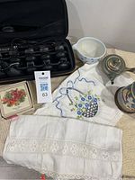 Topstyler hair waving system with clips laid out in open carrying case, surrounded by linens, a ceramic strawberry bowl, a small decorative ceramic container with lid, drink coasters box, and embroidered linens.