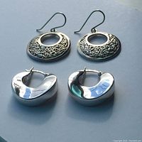 Two pairs of sterling silver earrings displayed on a flat surface: one pair round with patterned filigree and central hole with hook backs, one pair solid smooth thick half-hoops with latch backs