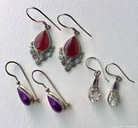 Photo showing three distinct pairs of sterling silver earrings: one with red stones and decorative details, one with purple stones, and one with clear stones, all with hooks or clips.
