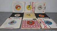 Overview of multiple 45 RPM vinyl records with sleeves from various artists and labels such as Columbia, PolyGram, and Warner-Elektra-Atlantic, arranged on a grey surface.