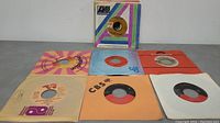 Photo showing six 45 RPM vinyl records in sleeves laid out on a table, featuring various label designs and colors.