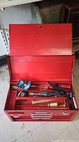 Opened red metal tool box showing various hand tools in the top compartment including a right angle pipe or wire threading tool, measuring tape, clamp, screwdrivers, and pliers.