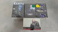 Four trading card sets displayed together on a gray surface: Battlefield Earth (still sealed box), Aliens vs. Predator (sealed), The Osbournes (sealed with visible pen marks on shrink wrap), and a single Game of Thrones pack with character art.