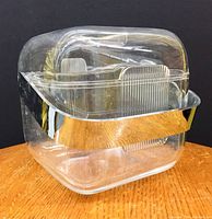 Clear lucite square ice bucket with rounded edges, removable lid, and silver-tone metal band handle, on wooden surface, front side view.