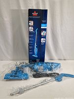 Photo showing vacuum parts laid out including the main vacuum unit, extension wand, power cord, and cleaning head wrapped in plastic, with the original Bissell Featherweight box behind