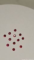 Thirteen small round faceted red rubies arranged on a flat white surface, showing their top facets and deep red color.