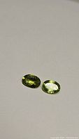 Two oval cut natural apple green peridot gemstones lying side by side on a light surface.