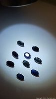 Nine dark blue oval cut sapphire gemstones arranged under a light spot, showing semi-transparent clarity and some inclusions.