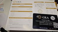Photo showing GRA grading certificate featuring detailed grading results for the moissanite, including cut, color, clarity, and carat weight with anti-counterfeit sticker and warranty card.