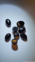 Eight polished black and chocolate onyx cabochon gemstones under direct light showing smooth oval and round shapes, with some stones displaying natural color variations and banding.