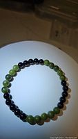 Photo shows a circular stretchy bracelet with alternating polished green jade beads and black onyx beads, each bead about 6mm in diameter, on a white surface under lighting.
