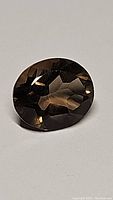 Oval cut natural smokey quartz gemstone on a plain background, showing deep brownish-gray color and faceted surface.