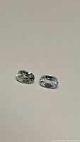 Two oval cut gemstones displayed on a plain background, showing off white to light blue coloration, likely Cambodian zircon.