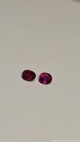 Two oval natural rubies placed side by side on a flat surface under normal lighting, showing bright pink-red color and clarity.