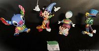 Four colorful Disney by Britto figurines featuring Stitch, Mickey Mouse Sorcerer's Apprentice, Cheshire Cat, and Jiminy Cricket, arranged side by side against a dark background.