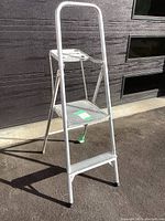 Front side view of entire aluminum folding step ladder standing on pavement, showing three steps including top step and handlebar.