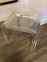 Three clear nesting tables stacked together, made of transparent acrylic, with curved edges, placed on dark wood flooring.