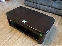 Dark wood rectangular coffee table on wheels, top surface shows scratches, visible sticker labels, rounded corners, lower shelf for storage.