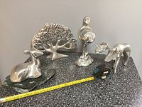 Overview of four aluminum sculptures on a speckled tabletop with measuring tape laid across
