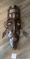 Photo showing large metal mask wall art lying on floor, presenting front face details and attached rings.