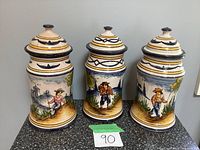 Front view of the three ceramic canisters with lids on, showing hand-painted countryside scenes with figures and decorative patterns.