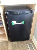 Front view of small black Danby refrigerator showing door and size comparison with surrounding cabinetry and floor vent.