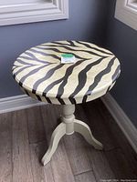 Front view of a round wooden table showing the zebra pattern on the top surface and sides with a turned pedestal base in cream color.
