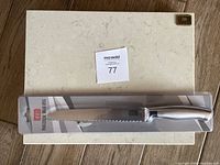 Photo shows Venoly professional bread knife still in original plastic packaging placed on top of a rectangular light beige Cambria stone cutting board with subtle marbled pattern and Cambria metal logo in top right corner.