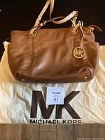 Front view of tan leather Michael Kors shoulder bag with MK logo charm and dust bag beneath.
