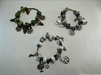 Photo of three charm bracelets laid flat on white surface, showing their beads and various metal charms including pentagrams, stars, horses, angel wings, and trees.