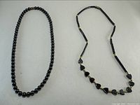 Two haematite necklaces laid flat on a light surface, one with round beads and one with mixed heart-shaped and cylindrical beads.
