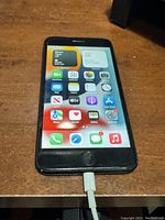 Front view of powered on iPhone 7 Plus showing home screen apps and charging cable connected.