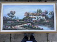 Full view of large oil painting with cottage, trees, water, flowers, and framed in white and gold frame.